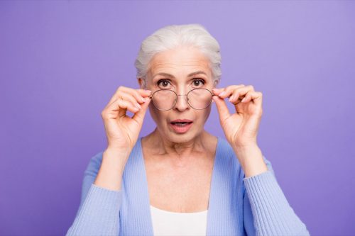 Portrait of beautiful nice amazed gray-haired old lady wearing casual and glasses, showing gesture, putting spectacles down. Isolated over pastel violet purple background