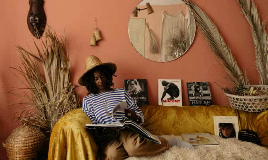 Maggie Coker wearing a straw hat, looking through a book sitting on her sofa, tall dried plants either side. The wall behind is burnt orange, the sofa has a mustard-coloured throw