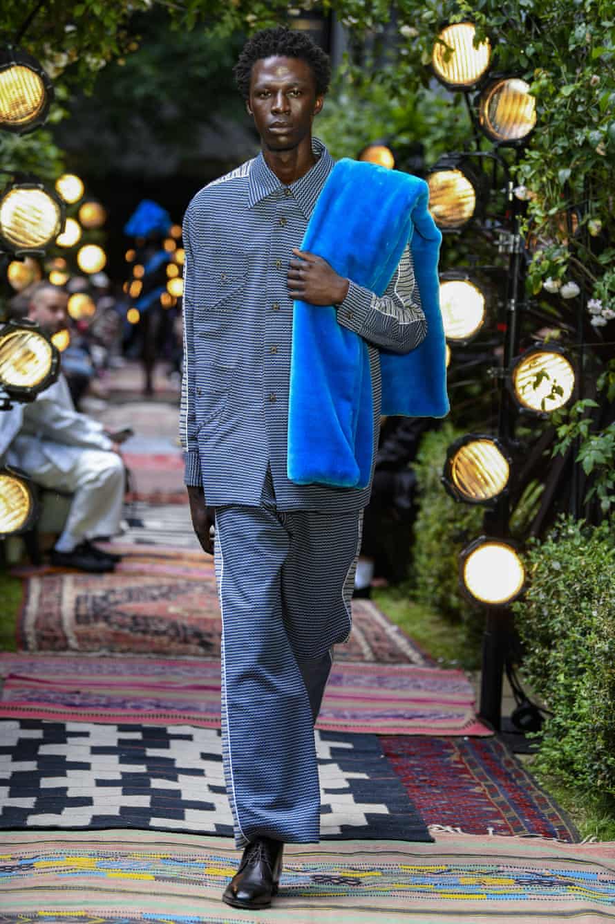 A Black male model is walking down the catwalk wearing a blue shirt and matching trousers, with a faux fur stole over his shoulder. 