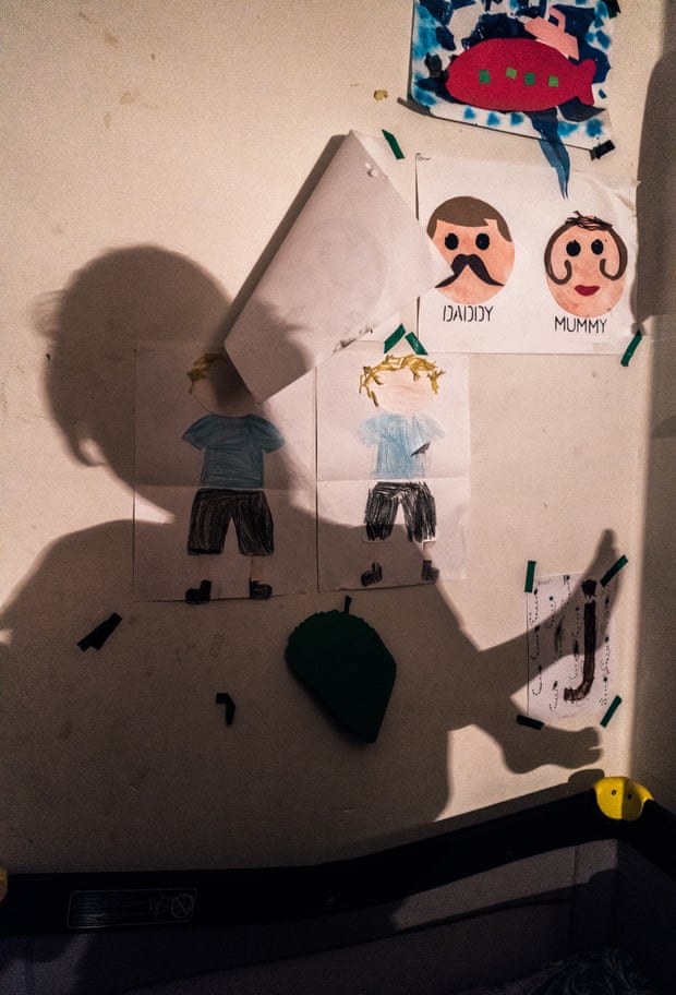 Some children’s drawings on a wall above a cot, with the shadow of a woman holding a child cast on the wall