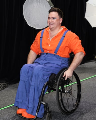 A man wears an orange shirt and shoes and blue overalls while sitting in a wheelchair