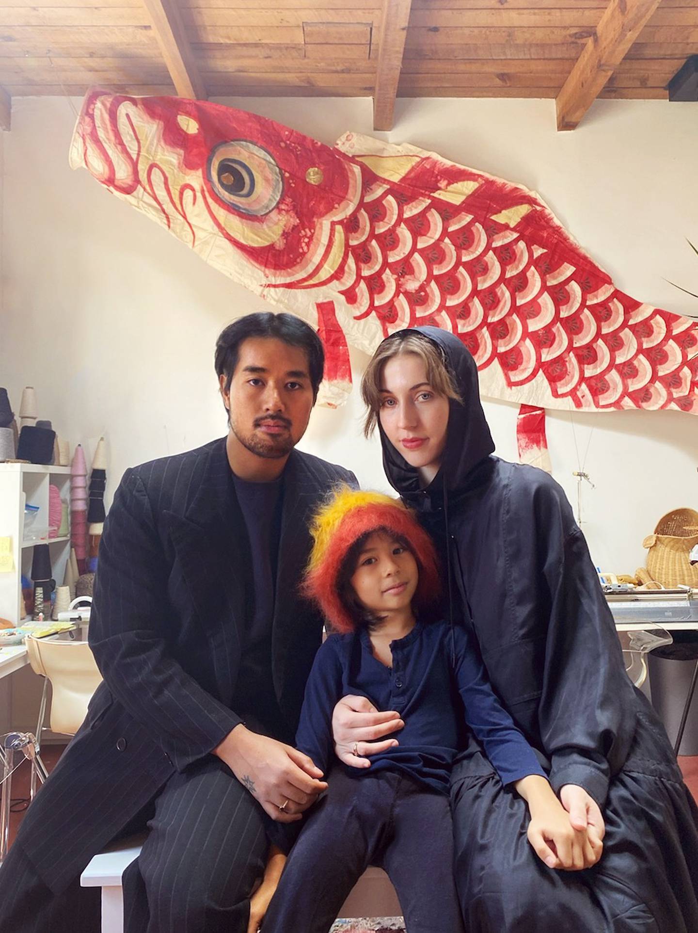 A man and woman sit on a table with a child. The child is wearing a fuzzy yellow and orange hat.