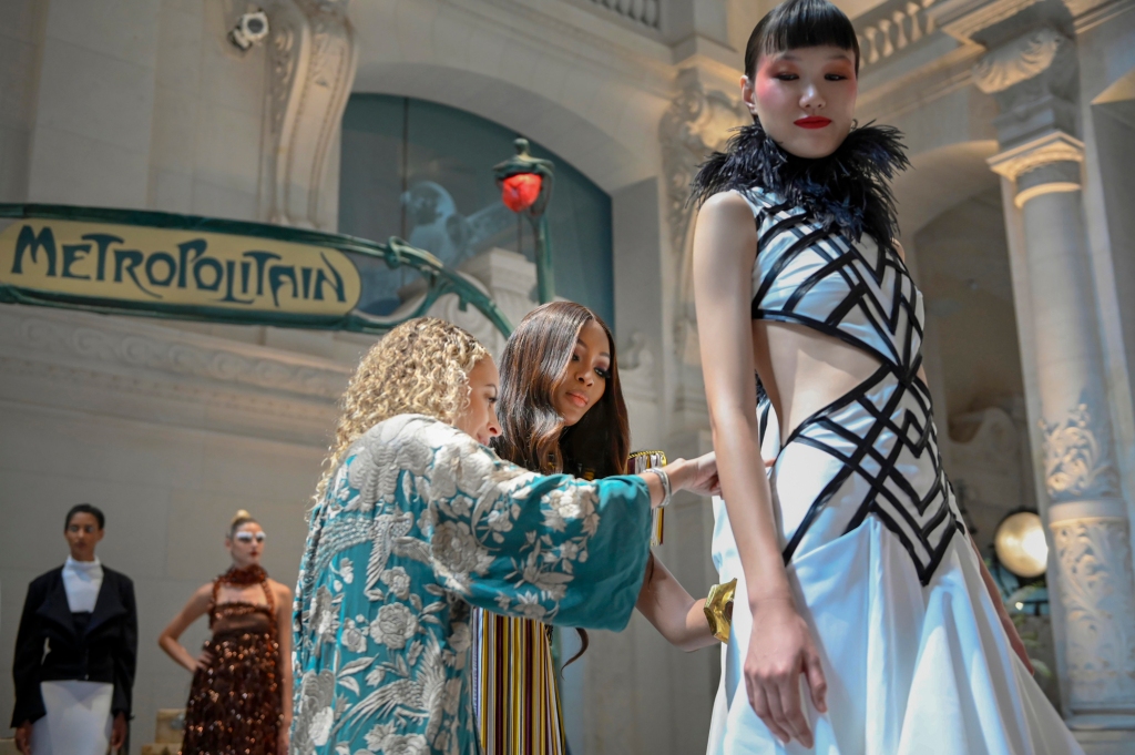 A model wears a dress while a fashion designer kneels and fusses with it. 