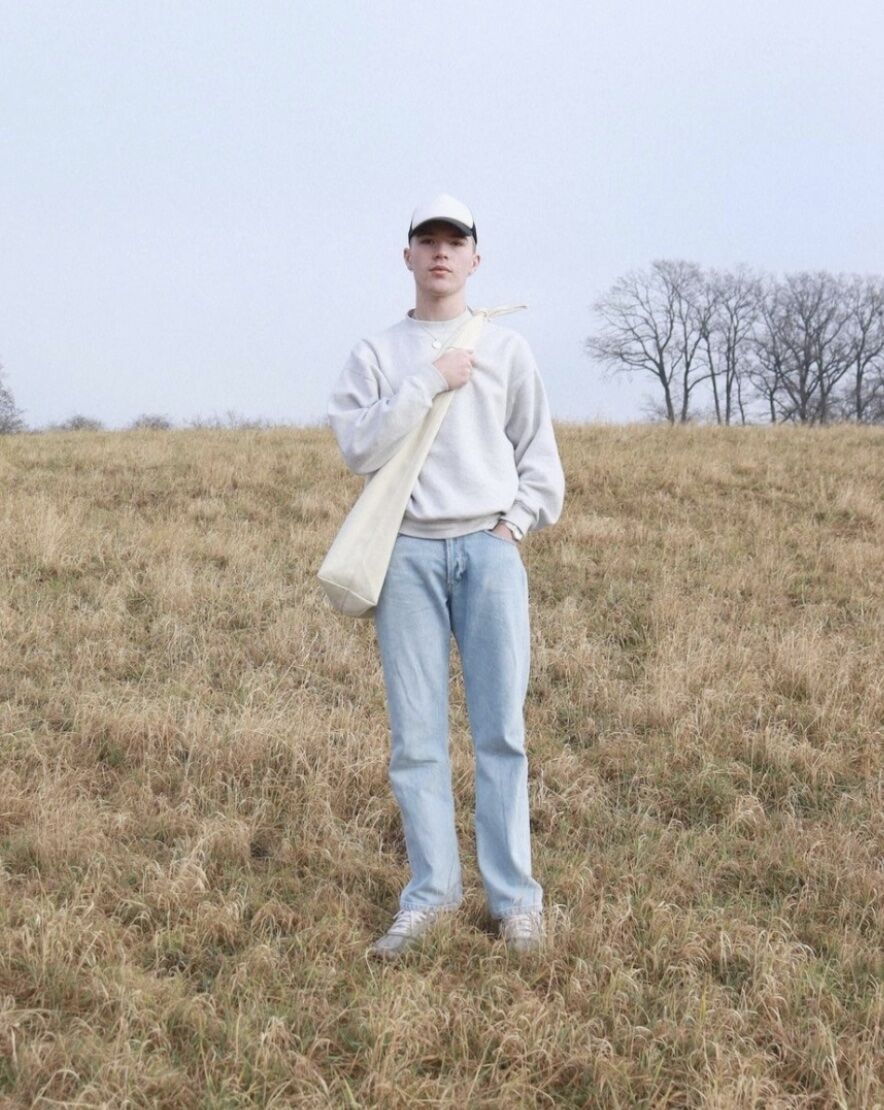 Man standing on hill wearing baseball hat, cross bag, and a sweater