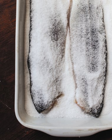 Two sea bass fillets are next to each other in a deep white dish, they are sitting on salt and sugar which is also covering the fillets