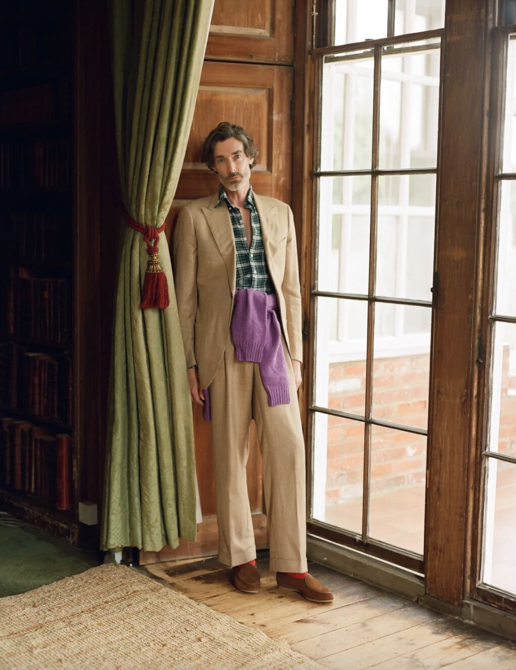 Man leaning against drapes wearing a casual suit and purple sweater around his waist