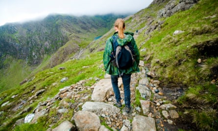 A woman standing on a rocky hiking trail, wearing a patterned green rain jacket and blue pants, with hiking boots and a backpack on. She looks out at a grassy green mountainside