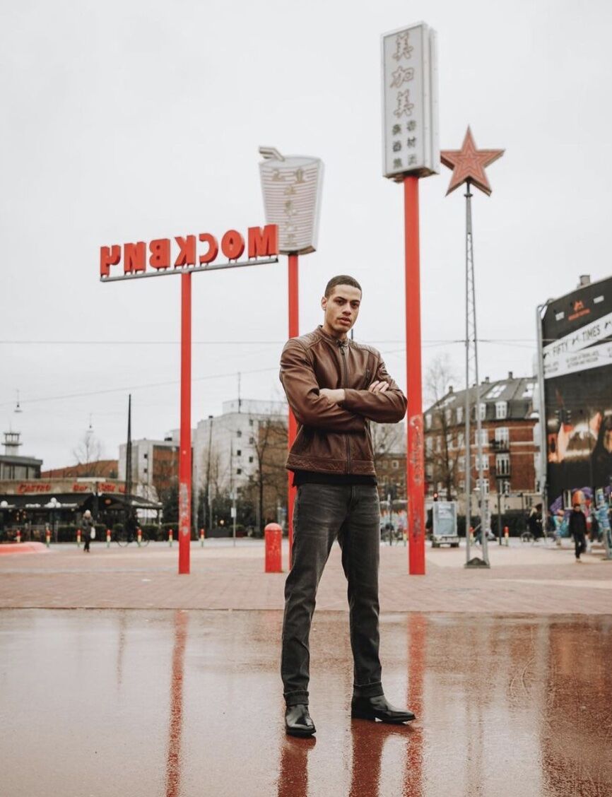man standing in front of large signs, arms crossed wearing a leather jacket, jeans and chelsea boots