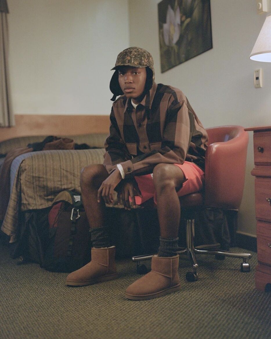 man sitting on a chair near a bed wearing a hat, shorts and UGG Boots