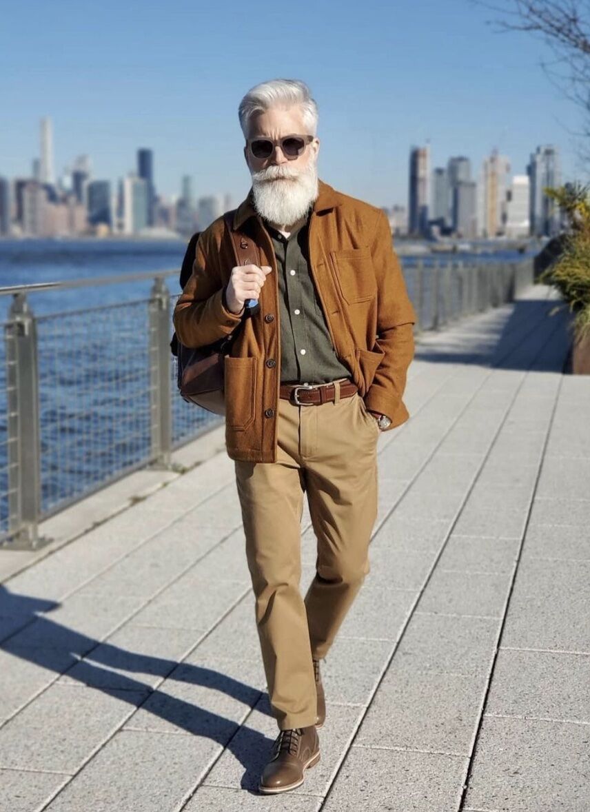 man walking on a boardwalk holding one strap of a backpack, groomed white beard and sunglasses 