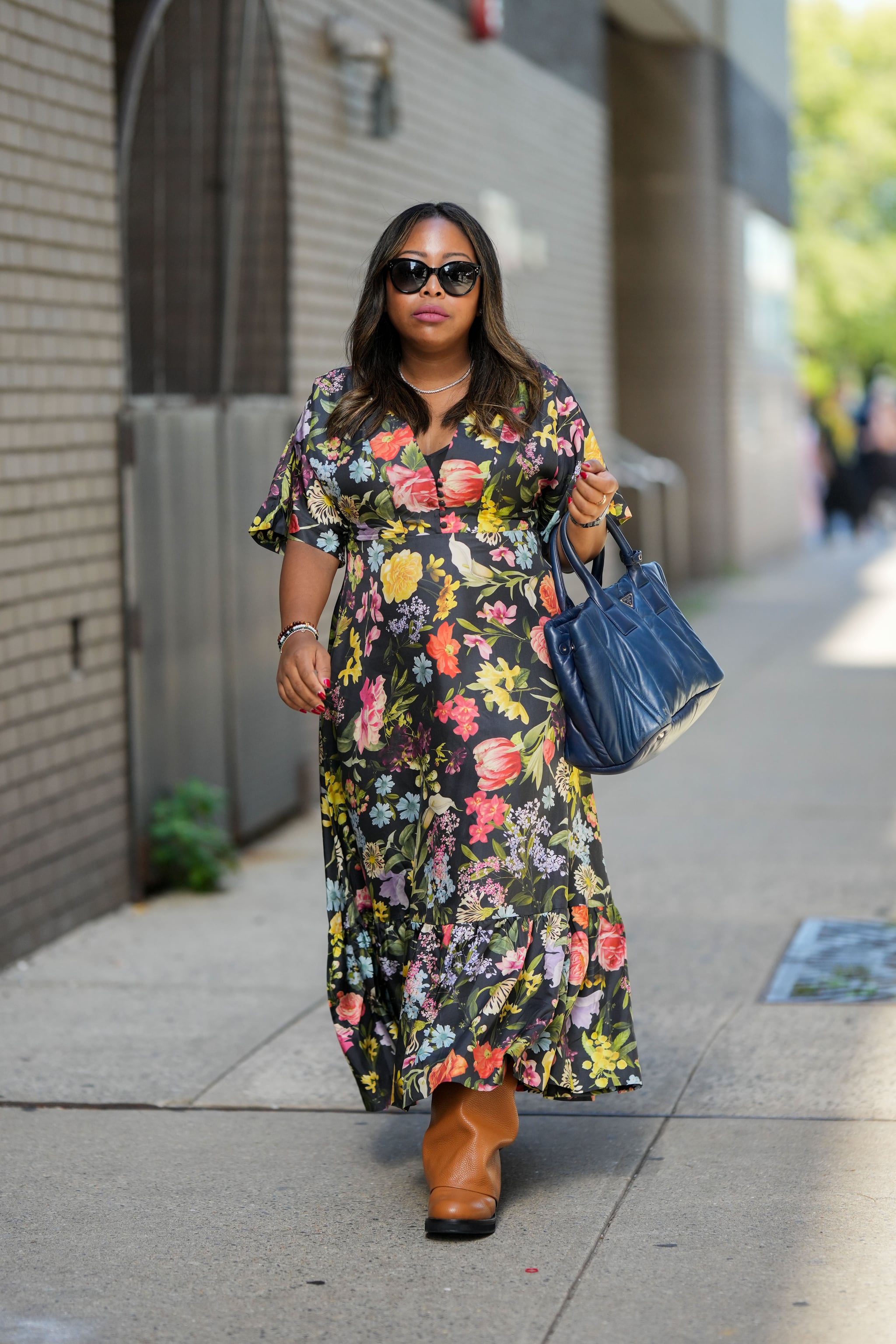 NEW YORK, NEW YORK - SEPTEMBER 10: A guest wears black sunglasses, a diamonds necklace, a navy blue shiny leather large handbag from Prada, gold rings, a silver watch, bracelets, a black with multicolored flower print pattern short sleeves / long dress, brown shiny leather knees boots , outside Altuzarra, during New York Fashion Week, on September 10, 2022 in New York City. (Photo by Edward Berthelot/Getty Images)