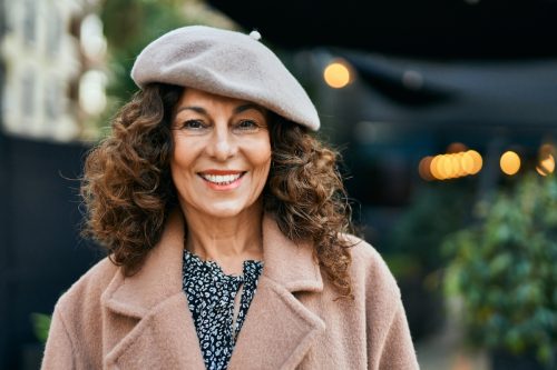 A middle-aged smiling woman with curly brown hair wearing a mauve peacoat and a gray beret.