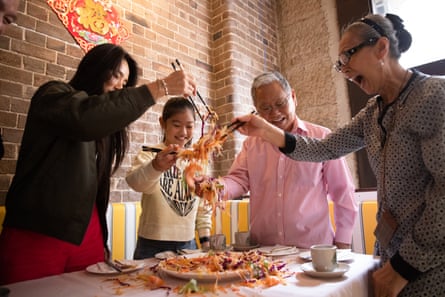 A multi-generational family stands at a table with chopsticks, tossing a salad of raw vegetables and salmon for lunar new year