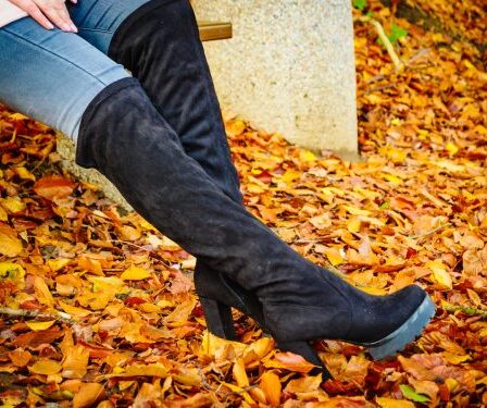 Close up of a woman's legs wearing jeans and black knee-high boots with orange leaves on the ground.