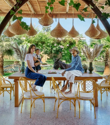 Cyrielle Rigot and Julien Tang and their son sit on a table in their outdoor dining room and cane lampshades hang above them, nearby is one of their donkeys in the outdoor dining room.