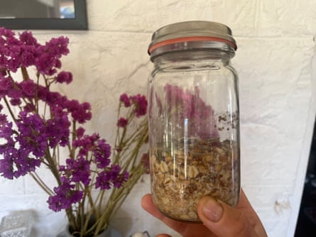 A hand holding a glass jar containing rolled oats and sunflower seeds against a white wall. A jar of tall purple flowers are in the background.