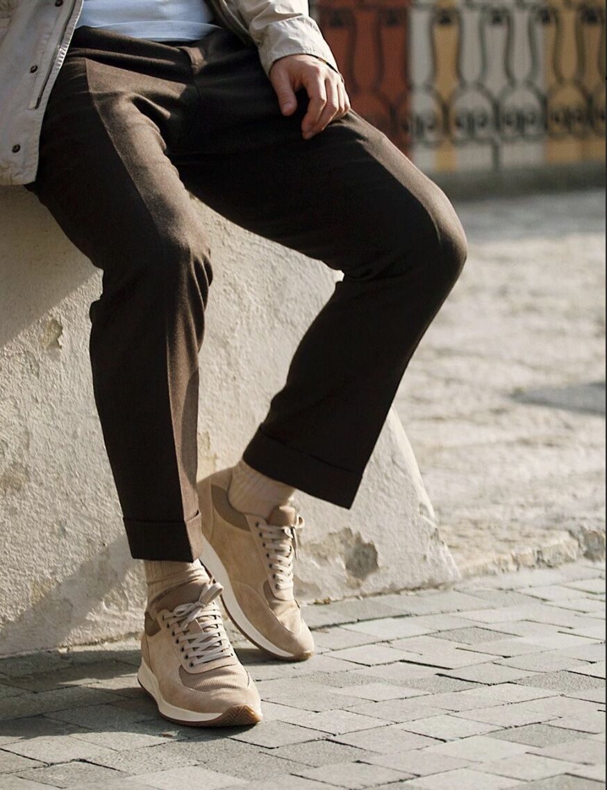 tailored brown trousers, tan socks and off white sneakers leaning against a wall outside