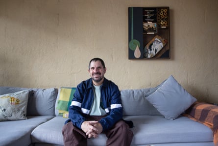 A man in a blue jacket sitting on a blue-grey couch. On the wall behind him is an interpretive portrait featuring skateboarding parts.