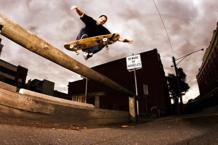 A wide-lens photograph of a man on a skateboarding leaping over a wood barrier against a cloudy sky.