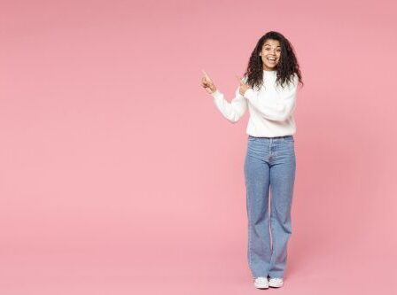 A smiling woman wearing a white turtleneck sweater, jeans, and white converse sneakers against a pink background