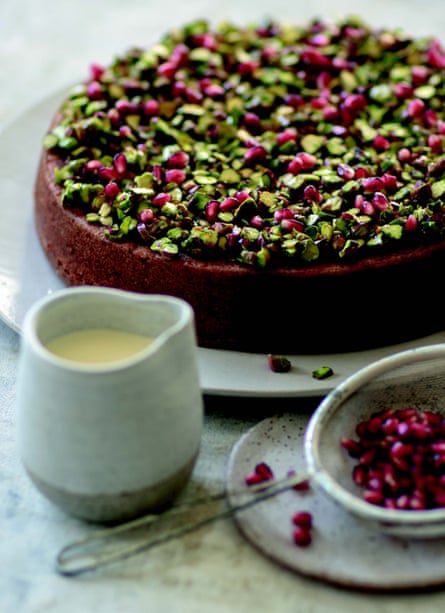 A pistachio and lime syrup cake decorated with chopped pistachios and pomegranate seeds, with a small jug of cream. 