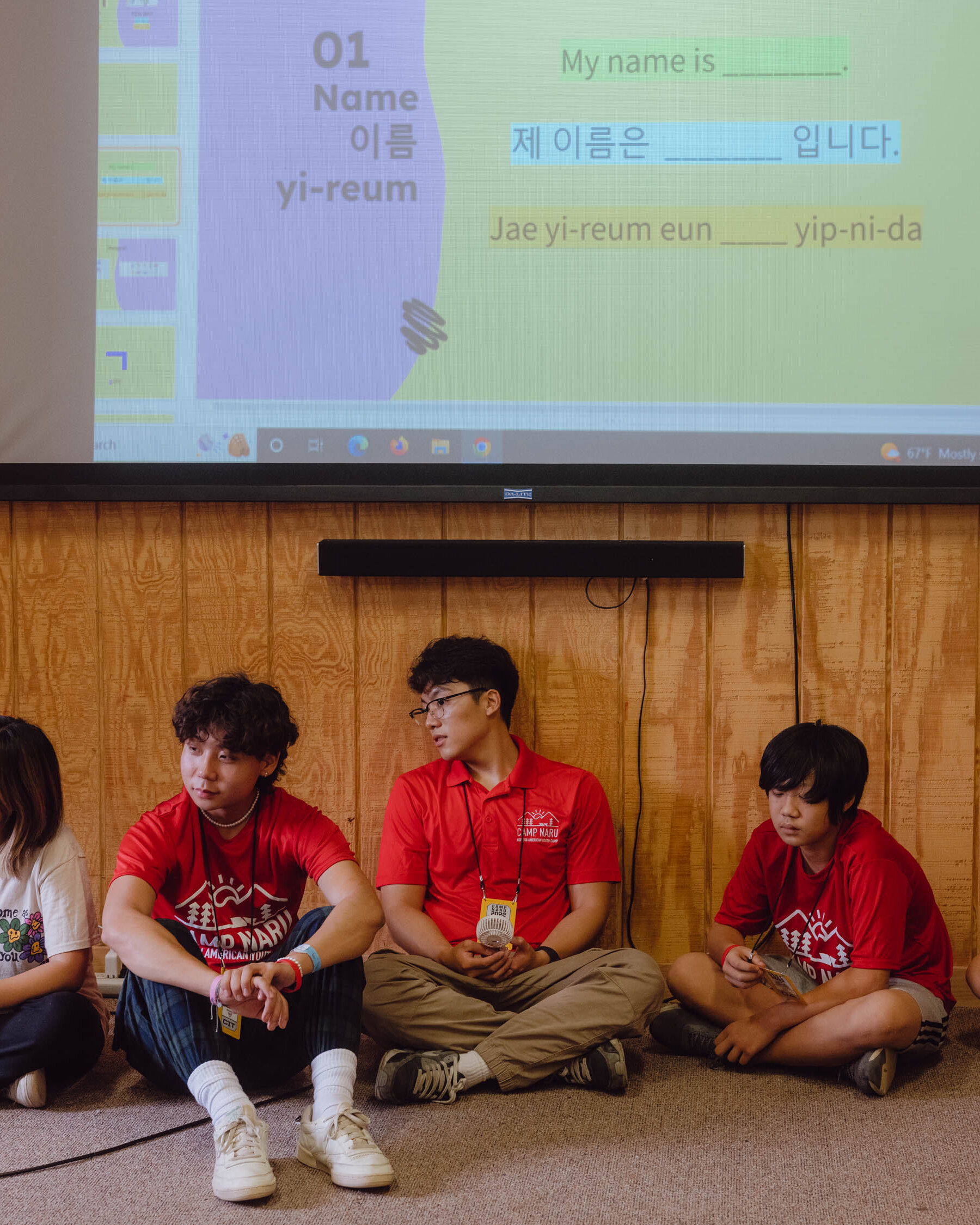 Five young people sit on a carpeted floor against a wood paneled wall, below a projector screen with an English and Korean lesson projected on it. Three of the children wear red T-shirts that say Camp Naru. The fourth, who is older, wears a red Camp Naru polo. The fifth, on the end, wears a white T-shirt with pink, green and yellow flowers on it.