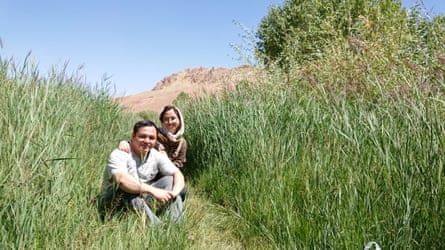 A young man, and a young woman in a hijab, in a field of tall grass, with mountains in the distant background