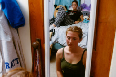 The reflection of two girls in a white-framed mirror, which is hanging over a wod door in a bedroom. The girl in front is sitting on the floor and wearing a dark green tank top. The girl in the back is reclining on a bed and wears a black T-shirt and plaid pajama pants.