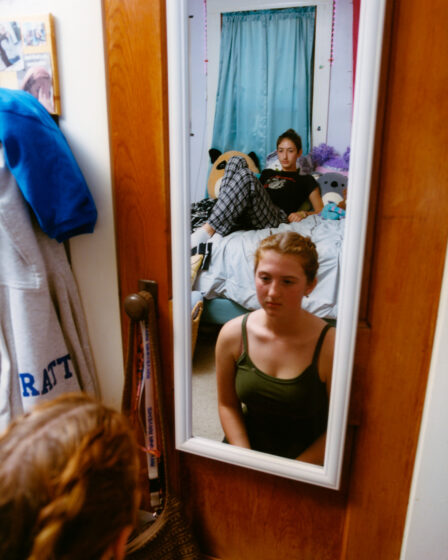 The reflection of two girls in a white-framed mirror, which is hanging over a wod door in a bedroom. The girl in front is sitting on the floor and wearing a dark green tank top. The girl in the back is reclining on a bed and wears a black T-shirt and plaid pajama pants.