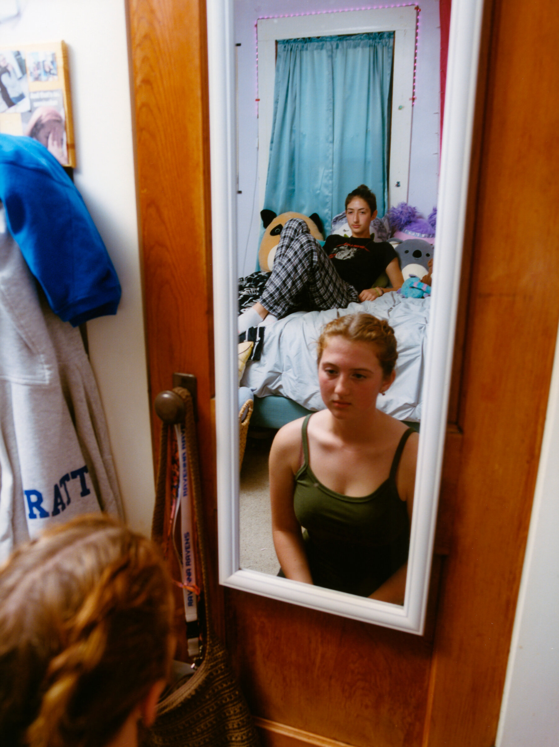 The reflection of two girls in a white-framed mirror, which is hanging over a wod door in a bedroom. The girl in front is sitting on the floor and wearing a dark green tank top. The girl in the back is reclining on a bed and wears a black T-shirt and plaid pajama pants.