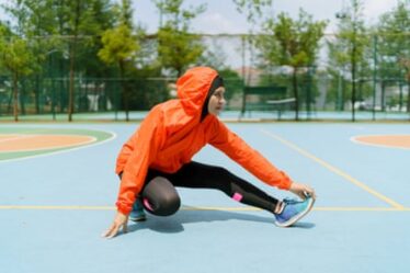 A woman in a hijab, orange hooded rain jacket, black tights and sports shoes, stretching her legs on a tennis court.