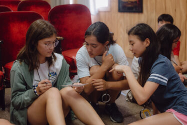 Three girls sitting on the floor of a wood-paneled room, in front of rows of red velvet seats. The girl on the left wears a white T-shirt, a green sweatshirt and glasses and holds a pen and a piece of paper. The girls on the right wear a white T-shirt and a blue T-shirt and pink shorts and gesture toward the piece of paper.
