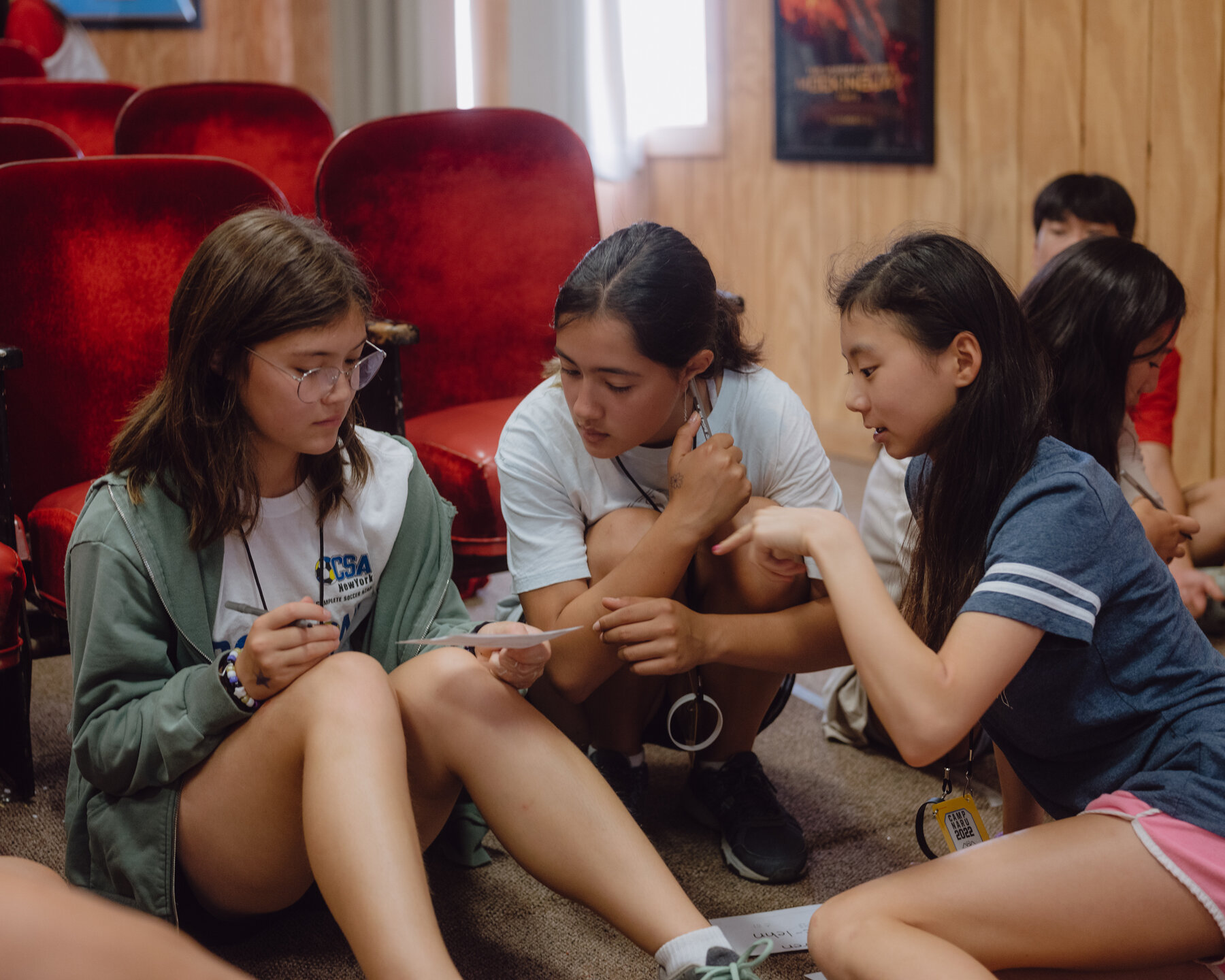 Three girls sitting on the floor of a wood-paneled room, in front of rows of red velvet seats. The girl on the left wears a white T-shirt, a green sweatshirt and glasses and holds a pen and a piece of paper. The girls on the right wear a white T-shirt and a blue T-shirt and pink shorts and gesture toward the piece of paper.