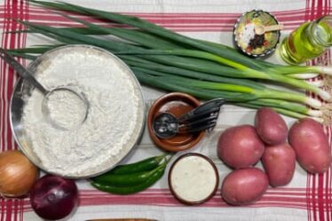 A bowl of flour, a bunch of spring onions, two onions, some green chillies, plus bowls of yoghurt and oil, laid out on a red and white tea towel