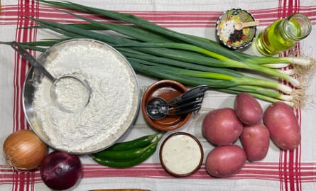 A bowl of flour, a bunch of spring onions, two onions, some green chillies, plus bowls of yoghurt and oil, laid out on a red and white tea towel