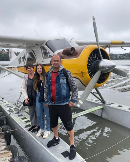 A mother, father and their adult daughter pose in front of a seaplane against a cloudy sky.