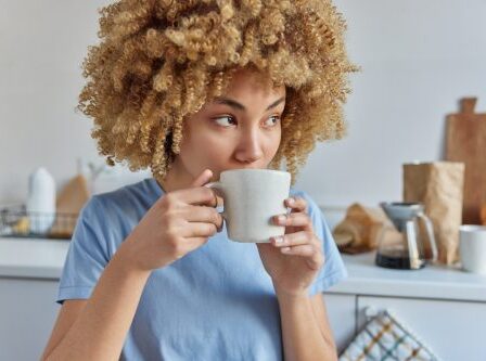 A young woman with curly hair wearing a sky-blue t-shirt drinks coffee in her kitchen