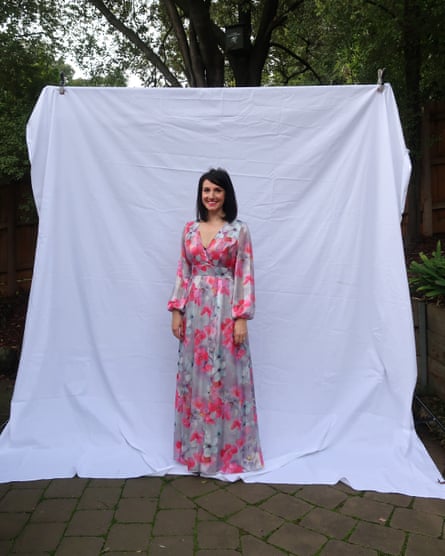 A woman in a floral pink long-sleeved dress stands in front of a white fabric backdrop in a leafy courtyard.