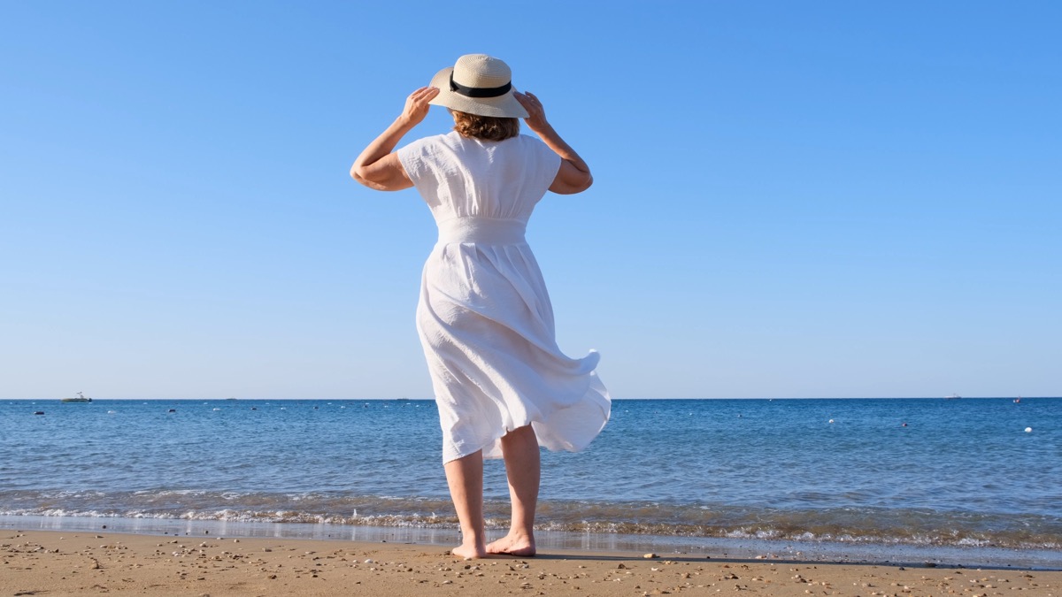 back view on a mature woman in a straw hat and white dress walks along the blue sea coast on a sunny summer day, enjoying freedom and relaxation. The concept of a typical life of pensioners.