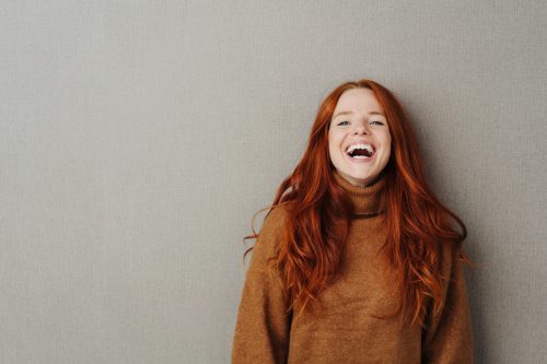 Laughing young redhead woman wearing a brown sweater with a vivacious smile posing over a grey studio background with copy space