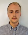 A man with short, light hair and a minimal beard looks at the camera with a plain expression. He is wearing a blue and white chequered shirt