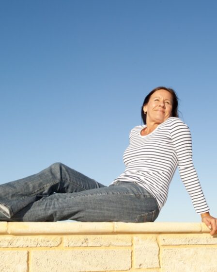 Portrait pretty mature woman sitting on a sunny day relaxed and happy on limestone wall, isolated with clear blue sky as background and copy space.