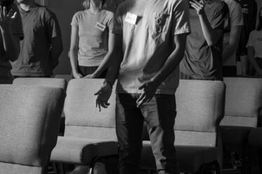 A group of students standing in rows of upholstered gray chairs in a chapel and singing. The young man at the front wears dirty white Converse sneakers, black pants, a light gray T-shirt and a white name tag that says