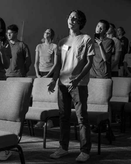 A group of students standing in rows of upholstered gray chairs in a chapel and singing. The young man at the front wears dirty white Converse sneakers, black pants, a light gray T-shirt and a white name tag that says