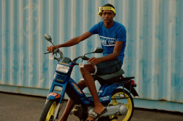 A young man sitting on a blue motorbike with yellow spokes. He wears a blue T-shirt that says