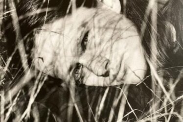 A black and white image of Nikki Brewster as a young woman, lying down in tall grass