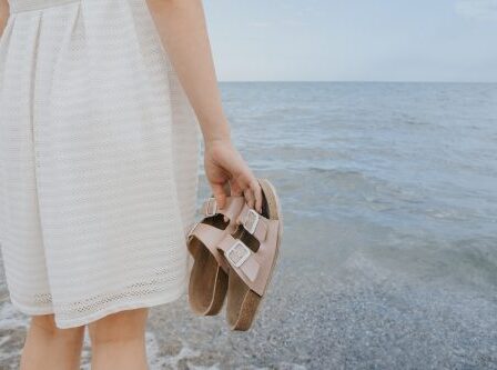 Woman in an off-white dress stands near the ocean holding beige Birkenstock sandals