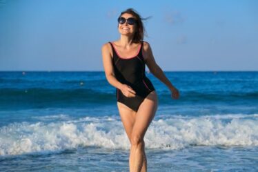 A smiling woman walking out of the ocean wearing a sporty black one-piece bathing suit and sunglasses