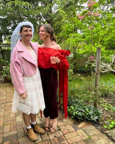 A man dressed in a wedding veil, white dress, pink jacket and boots, standing with a woman in a red blouse and black trousers