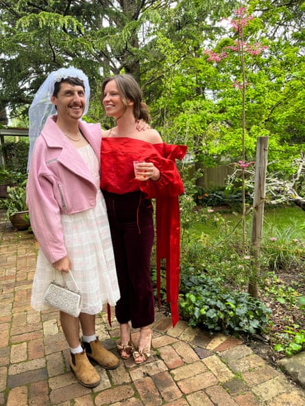 A man dressed in a wedding veil, white dress, pink jacket and boots, standing with a woman in a red blouse and black trousers